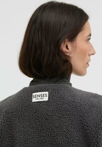 Gray fleece jacket with a textured surface and high collar, featuring a white label reading "SENSES .THE LABEL" on the back.