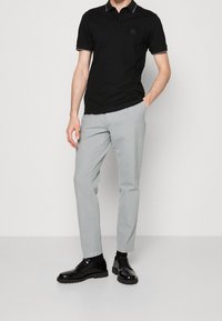 Homme portant un polo noir, un pantalon gris clair, des chaussettes noires et des chaussures noires à lacets, debout avec une main dans la poche.