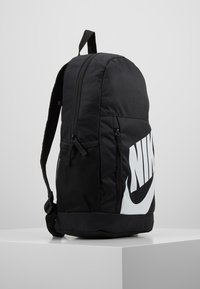 Sac à dos noir en tissu durable, avec un grand logo Nike blanc, des poches zippées et un panneau latéral en maille pour la ventilation.
