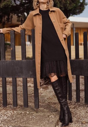 Kobieta opierająca się o czarny drewniany płot, ubrana w camelowy płaszcz, czarną sukienkę z koronkowym wykończeniem, podszewkę w panterkę oraz czarne kozaki za kolano na zewnątrz.