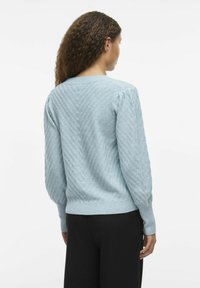 Lyseblå strikket genser med et strukturert sikk-sakk-mønster, rund hals, og litt puffede ermer, kombinert med svarte bukser.