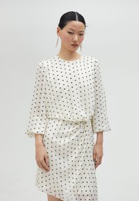 Vestido blanco con lunares negros, tela fluida, mangas largas tipo campana, detalle de cintura anudada y dobladillo asimétrico.