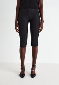 Black ribbed capri pants with side slits, featuring a fitted design. Worn with black pointed-toe heels with silver chain accents.