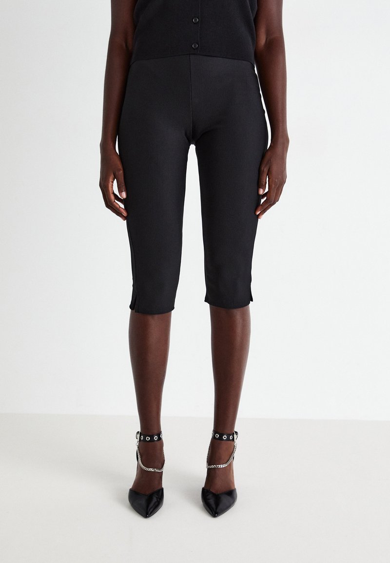Black ribbed capri pants with side slits, featuring a fitted design. Worn with black pointed-toe heels with silver chain accents.