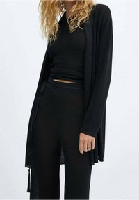 Cardigan en tricot noir à l'avant ouvert, associé à un haut côtelé et un pantalon assortis. Texture douce, coupe décontractée et design dépouillé.