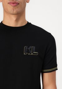 Juodas trumpas rankovės marškinėliai su apvaliu iškirpimu, puikuojantys blizgia KL logotipu juodai geltonais akcentais ir dryžuotomis auksinėmis detalėmis ant rankovių.