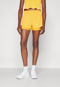 Gelbe Sportshorts mit einem elastischen Bund und einer kontrastierenden Innenauskleidung. Kombiniert mit weißen Sneakern und Kniestrümpfen.