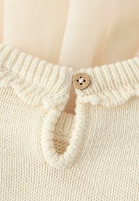 Kremfarget strikket plagg med bølget halslinning og en liten treknapp. Har en myk tekstur og subtil sømdekorasjon.