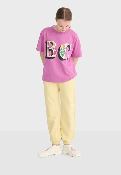 Jeune personne regardant vers le bas, portant un t-shirt rose avec des lettres florales "BC", un pantalon de survêtement jaune et des baskets blanches sur un fond uni.