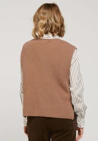 Gilet en tricot marron avec une finition texturée, présentant un décolleté rond et une couverture des épaules, porté par-dessus une chemise à manches longues rayée.