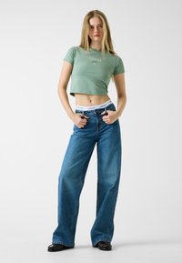 Zielona krótka koszulka z logo "GUESS", zestawiona z luźnymi niebieskimi jeansami o wysokim stanie i prostych nogawkach.