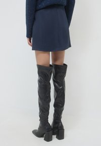 Donna in stivali neri al ginocchio con tacco e una gonna blu navy, in piedi contro uno sfondo chiaro uniforme, vista da dietro.
