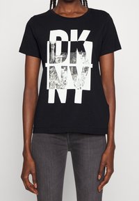 Svart bomullströja med en vit grafik som har "DKNY"-text och ett mönstrat bakgrund. Korta ärmar, avslappnad passform och sömlös halsringning.