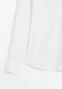 Chemise blanche à manches longues avec poignets et boutons visibles sur le devant, sur un fond blanc uni.