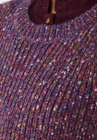 Maglione lavorato a maglia di colore viola con macchie multicolori. Presenta un motivo a coste e un colletto di velluto a coste color marrone scuro con un bottone dorato.