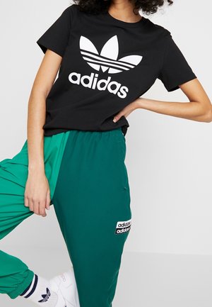 Osoba nosząca czarną koszulkę Adidas z białym logo, turkusowe spodnie Adidas z naszywką z logo, białe skarpetki Adidas oraz białe buty sportowe.