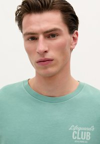 Muntgroene katoenen t-shirt met een ronde hals en korte mouwen. Bevat witte opdruk met de tekst "Lifeguard's CLUB" op de linkerborst.