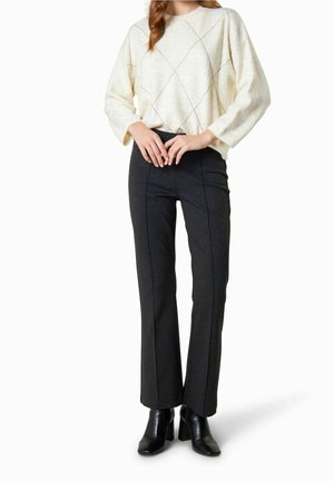 Mujer con suéter crema de patrón de diamantes, pantalones oscuros ajustados con costuras frontales y botines negros con tacón, posando frente a un fondo blanco.