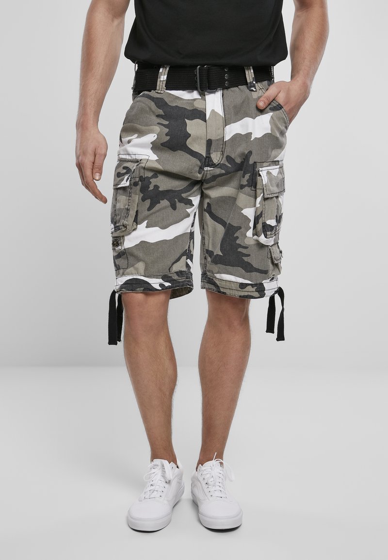 Homme portant un short cargo camouflage gris et noir avec plusieurs poches, une ceinture noire, des baskets blanches et un t-shirt noir avec une main dans la poche.