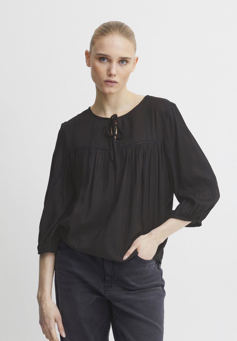 Blusa nera con scollatura rotonda, dettaglio a fiocco davanti e maniche a 3/4. Tessuto strutturato e design morbido. Abbinata a jeans scuri.
