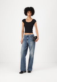 Svart kort topp med scalloped kanter, kombinerad med ljusblå vida jeans. Modellen håller en liten svart väska och bär svarta skor.