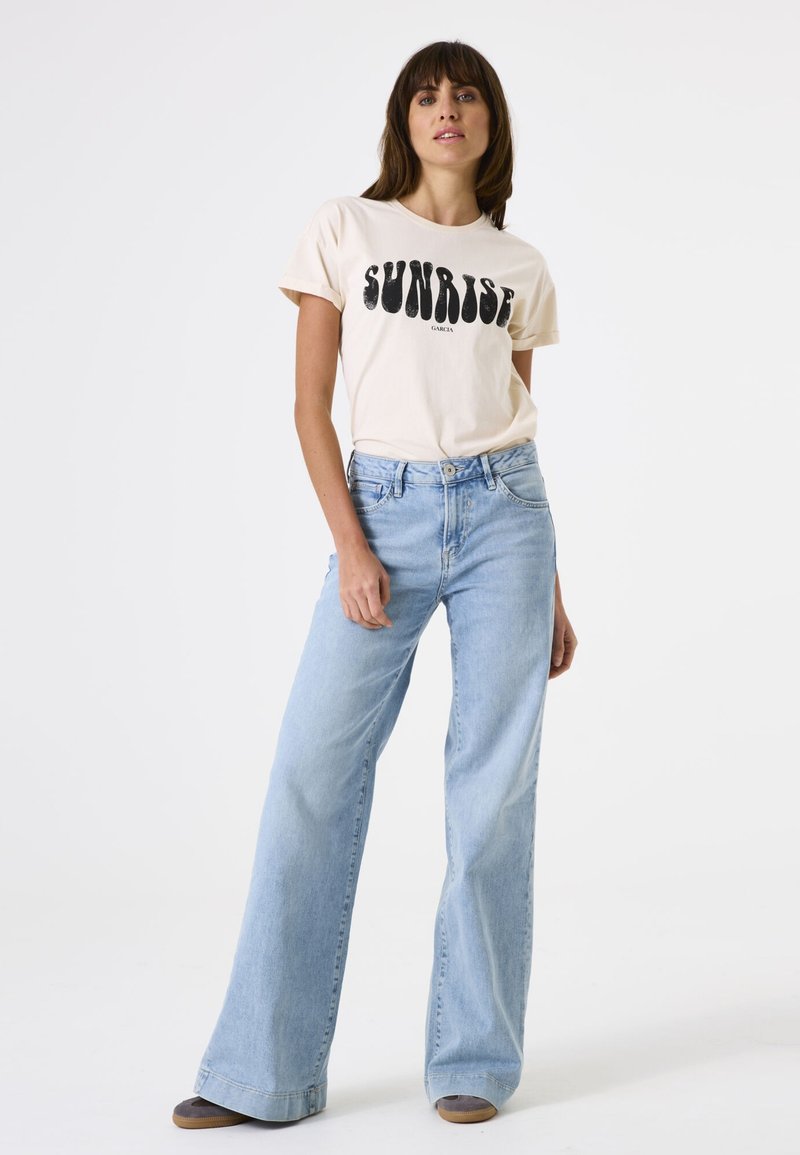 Hellbeige T-Shirt mit schwarzem "SUNRISE"-Grafik, kombiniert mit hochgeschnittenen, weit geschnittenen hellblauen Jeans. Weicher Stoff und lockere Passform.