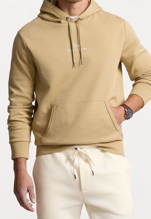 Muž nosící béžovou mikinu s kapsou vpředu a bílým nápisem „Polo Ralph Lauren“, doplněnou krémovými kalhotami se stahovací šňůrkou a náramkovými hodinkami.