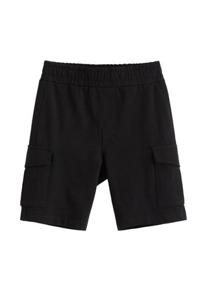 Smilende barn med krøllet blondt hår iført en grønblå trøje med et abeansigt, sorte shorts, grønblå sokker og hvide sneakers.