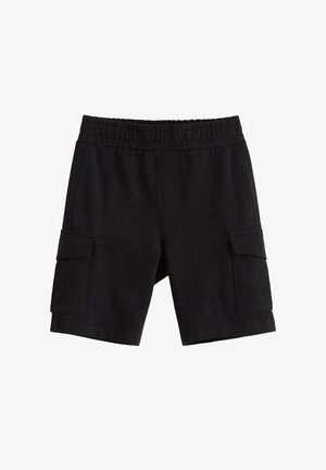 Smilende barn med krøllet blondt hår iført en grønblå trøje med et abeansigt, sorte shorts, grønblå sokker og hvide sneakers.