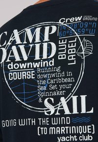 Marine blauwe katoenen T-shirt met witte en blauwe grafische tekst, nautische thema's en ronde designelementen, waarbij zeilcoördinaten worden benadrukt.