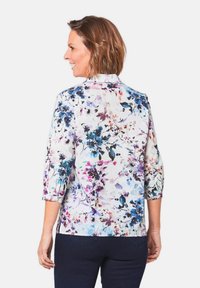 Blumenbluse mit 3/4-Ärmeln, aus leichtem Stoff mit einem weißen Grund und lebhaften lila, blauen und grünen Blumenmustern.