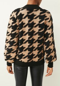 Sweter w czarno-beżowy wzór w jodełkę, wykonany z miękkiego, puszystego materiału, z prążkowanym czarnym dekoltem i mankietami. Widoczne są również czarne spodnie z ekoskóry.