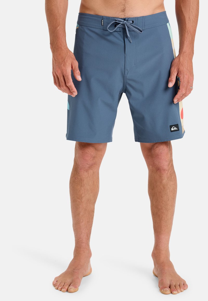 Pantaloni scurți albaștri din material ușor, cu talie ajustabilă prin șnur, având un model geometric multicolor pe părțile laterale și o mică etichetă cu logo pe margine.