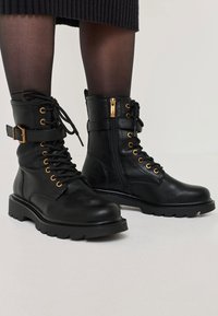 Stivali alla caviglia in pelle nera con hardware dorato, lacci frontali e zip laterale. Presentano una suola spessa e una cintura regolabile.