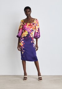 Blumenmuster-Kleid aus lila Satin mit puffigen Ärmeln, quadratischem Ausschnitt und knielanger Schnittführung. Modell trägt Sandalen mit Blockabsatz.