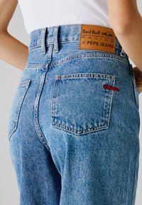 Jeans de mezclilla de color azul claro con dos bolsillos traseros, una etiqueta de cuero que dice "Red Bull Racing x Pepe Jeans" y bordado en rojo "RBR" en el bolsillo.