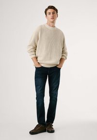 Beige gestrickter Pullover mit Rippenstruktur, kombiniert mit dunkelblauen Jeans und braunen Schnürschuhen. Stehende Pose mit Händen in den Taschen.
