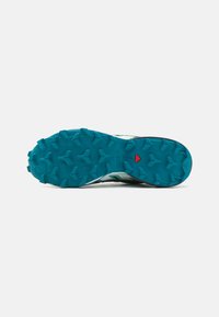 Semelle de chaussure de trail en teal et noir avec des crampons multidirectionnels pour une meilleure traction. Présente un accent rouge dans la zone du talon.