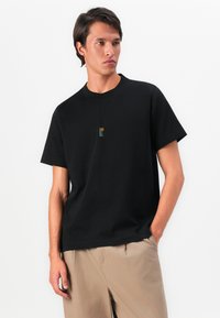 Bărbat tânăr îmbrăcat cu un tricou negru pe care se află o mică grafică cu un palmier și soare, asortat cu pantaloni bej, stând în fața unui fundal alb.
