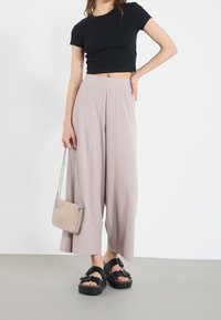 Černý žebrovaný crop top, světle růžové široké kalhoty s vertikální texturou, černé sandály na platformě a malá béžová kabelka se stříbrnými detaily.