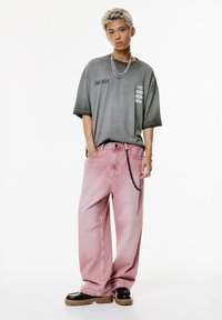 Ung mand med afbleget hår iført en grå oversized T-shirt, lyserøde vidde bukser med kæde og klodsede brune sko på en hvid baggrund.