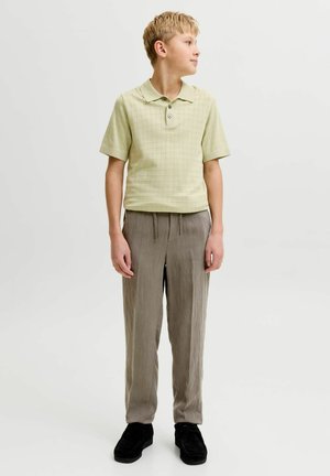 Plavi dječak stoji, nosi svijetlozeleno polo majicu kratkih rukava, široke bež hlače i crne cipele, gleda prema desno.