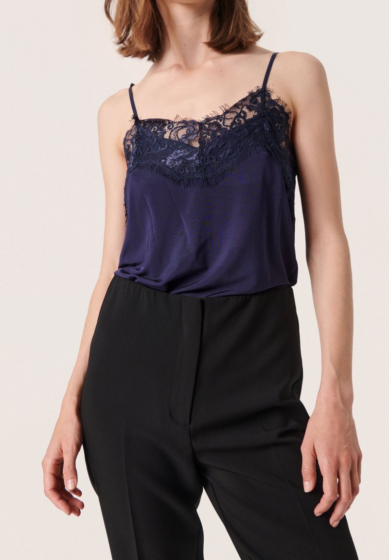 Camisola in satin blu navy con bordo in pizzo e spalline sottili, abbinata a pantaloni neri a vita alta. Texture liscia e dettagli in pizzo delicato.