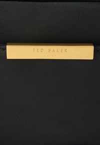 Μαύρη τσάντα με υφή, με μια πλακέτα σε χρυσό τόνο με ανάγλυφη την επιγραφή "TED BAKER LONDON." Ο σχεδιασμός είναι κομψός και μινιμαλιστικός.