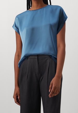 Personne portant un chemisier bleu à manches courtes rentré dans un pantalon plissé gris foncé à taille haute, debout devant un fond clair.