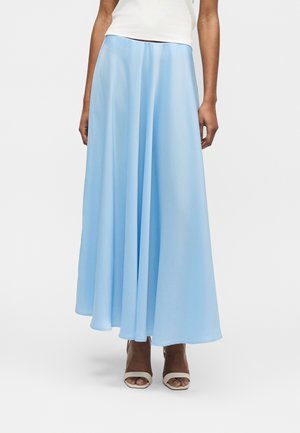 Mujer que lleva una falda larga azul claro fluida, camiseta blanca y sandalias beige de tacón con los dedos abiertos, de pie sobre un fondo blanco.