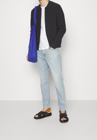 Chaqueta bomber negra, camiseta blanca, jeans azul claro y sandalias negras con acentos naranjas. Bolsa de tote azul colgada del hombro.
