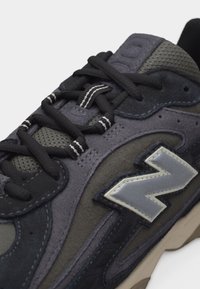 Μαύρο και γκρι αθλητικό παπούτσι με πάνελ από δίχτυ και σουέντ, μαύρα κορδόνια με λευκές ρίγες και αντανακλαστικό γκρι λογότυπο "N" στη πλευρά.