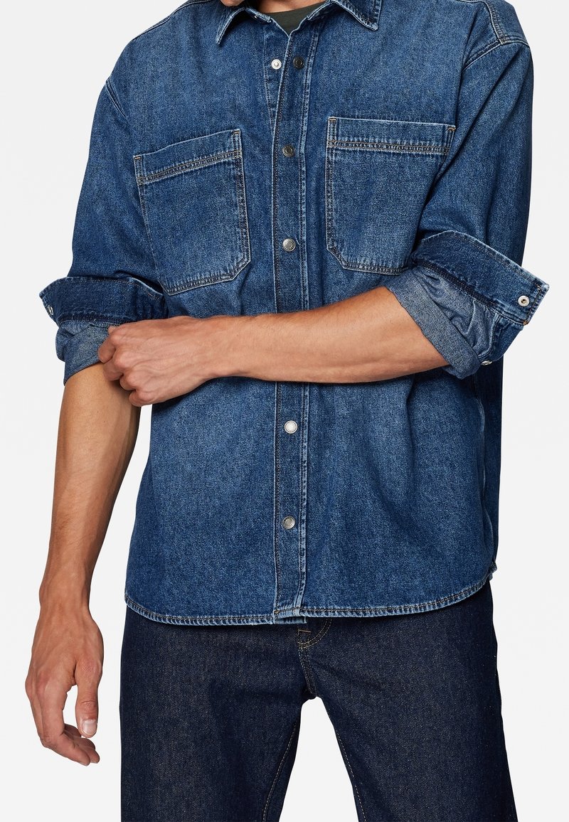 Denim overhemd in donkerblauw met knoopsluiting aan de voorkant, twee borstzakken en opgerolde mouwen. Voorzien van een klassiek kraag en een casual pasvorm.