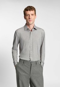Camicia grigia a maniche lunghe, con texture liscia, colletto classico e apertura con bottoni; abbinata a pantaloni grigi con tasche laterali.